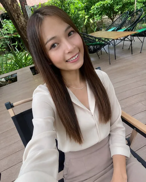 Smiling young woman with long brown hair on a wooden outdoor patio, wearing a white blouse and beige skirt amid lush greenery.