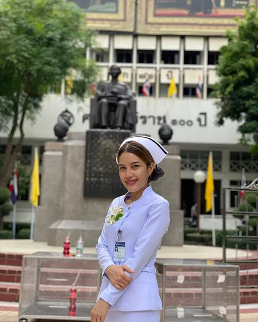 Smiling nurse in a white uniform and cap poses before a memorial statue outside a Thai hospital, surrounded by trees and yellow flags.